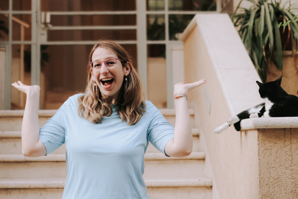 Tamara laughing with her arms wide open on a staircase, a black and white cat lying next to her