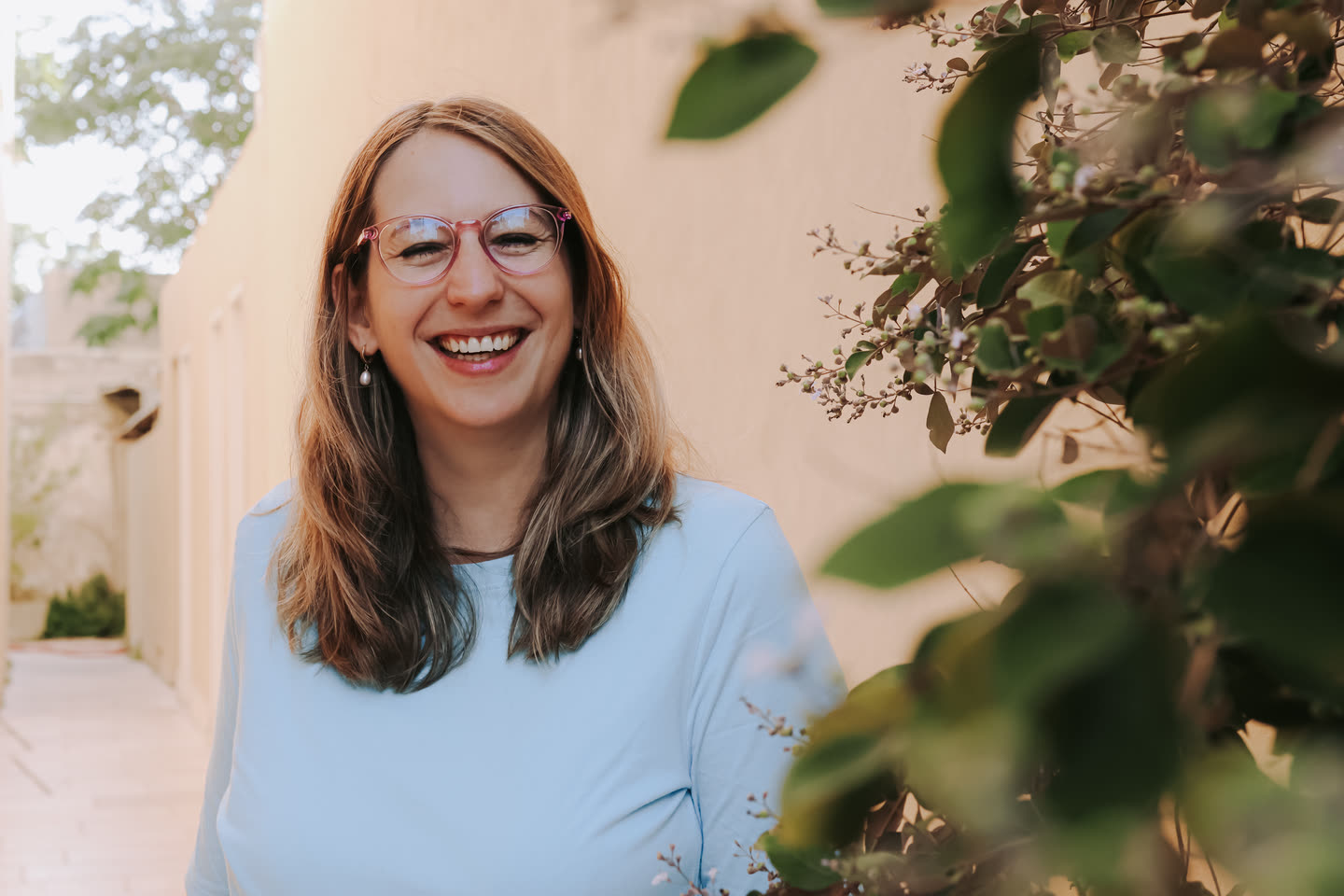 Tamara smiling at the camera in a light blue t-shirt, green leaves beside her