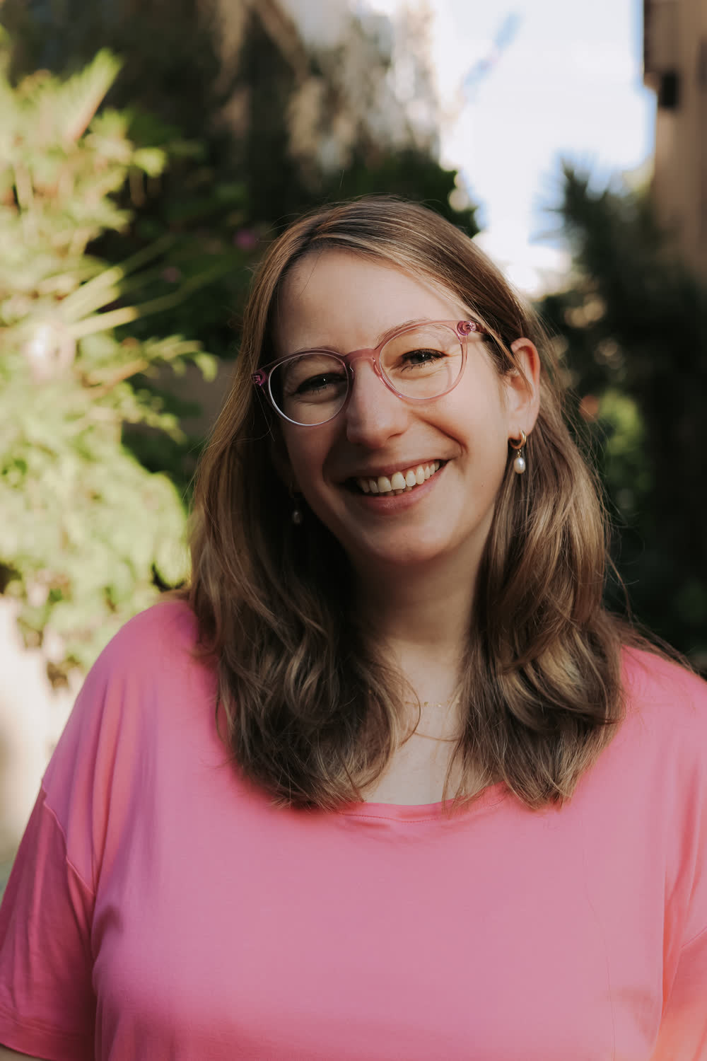 Tamara smiling at the camera in a pink t-shirt, green leaves behind her