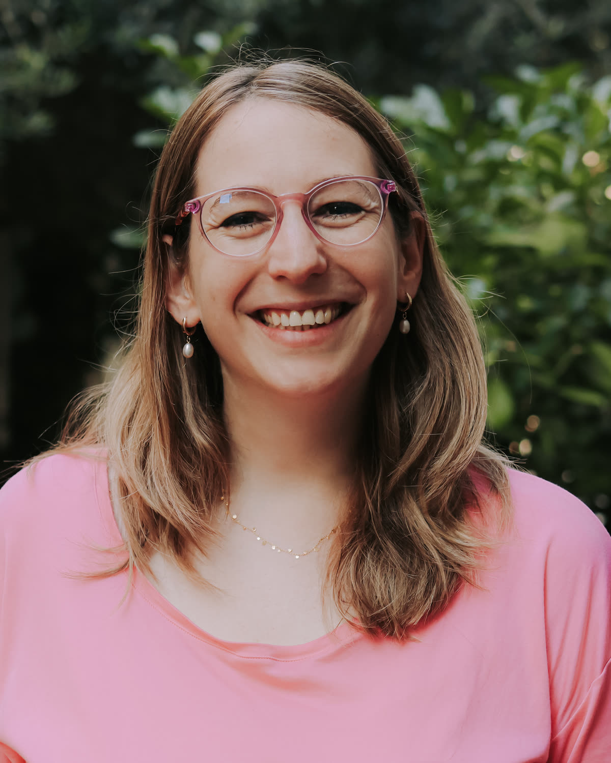 Tamara smiling at the camera in a pink t-shirt, green leaves behind her