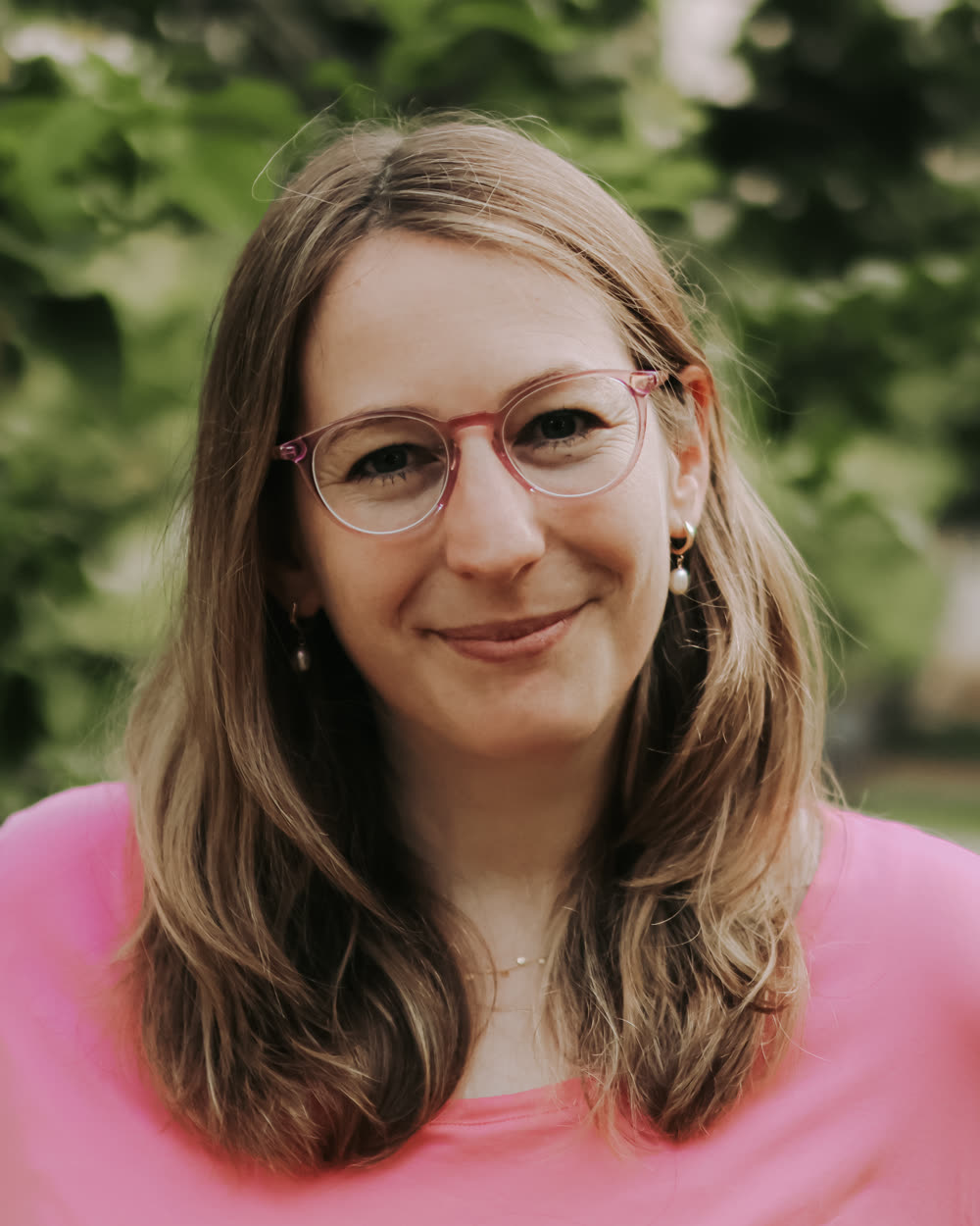 Tamara smiling at the camera in a pink t-shirt, green leaves behind her